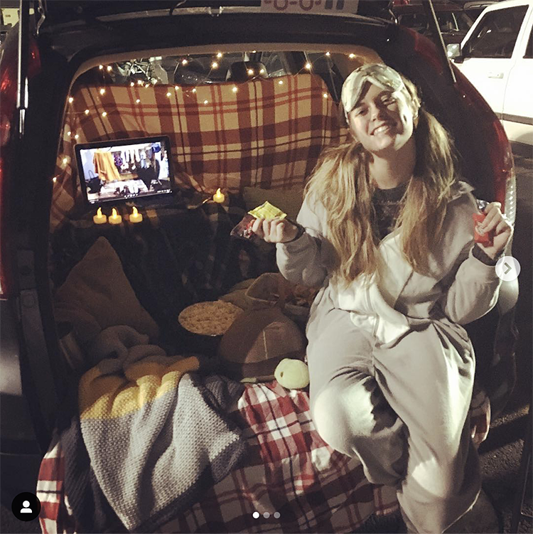 A trunk decorated with pajama party items including candles, a movie screen and junk food. A person is dressed in pajamas and a face mask. This is an easy trunk or treat Halloween decorating idea.