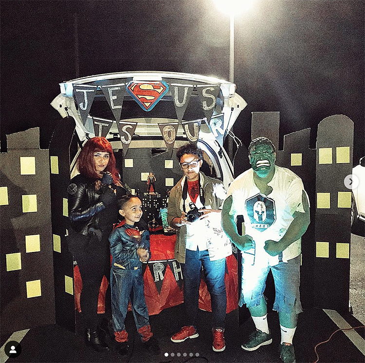 Trunk decorated as a city from a superhero movie. A sign reads "Jesus is our Superhero". The family is dressed as various superhero characters to match the trunk or treat theme. | The Dating Divas