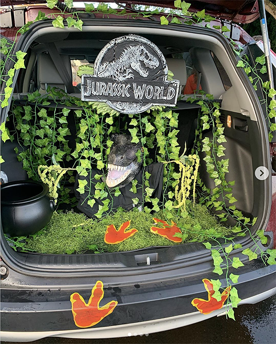 Trunk decorated with a theme for the movie Jurassic Park. It includes a lot of vines and greenery, the park's logo and printed dinosaur footprints leading to the trunk. This is a great trunk or treat idea | The Dating Divas