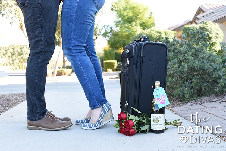 Couple has a bag packed, roses, sparkling cider and shares a kiss as they prepare for their romantic hotel getaway anniversary ideas| The Dating Divas