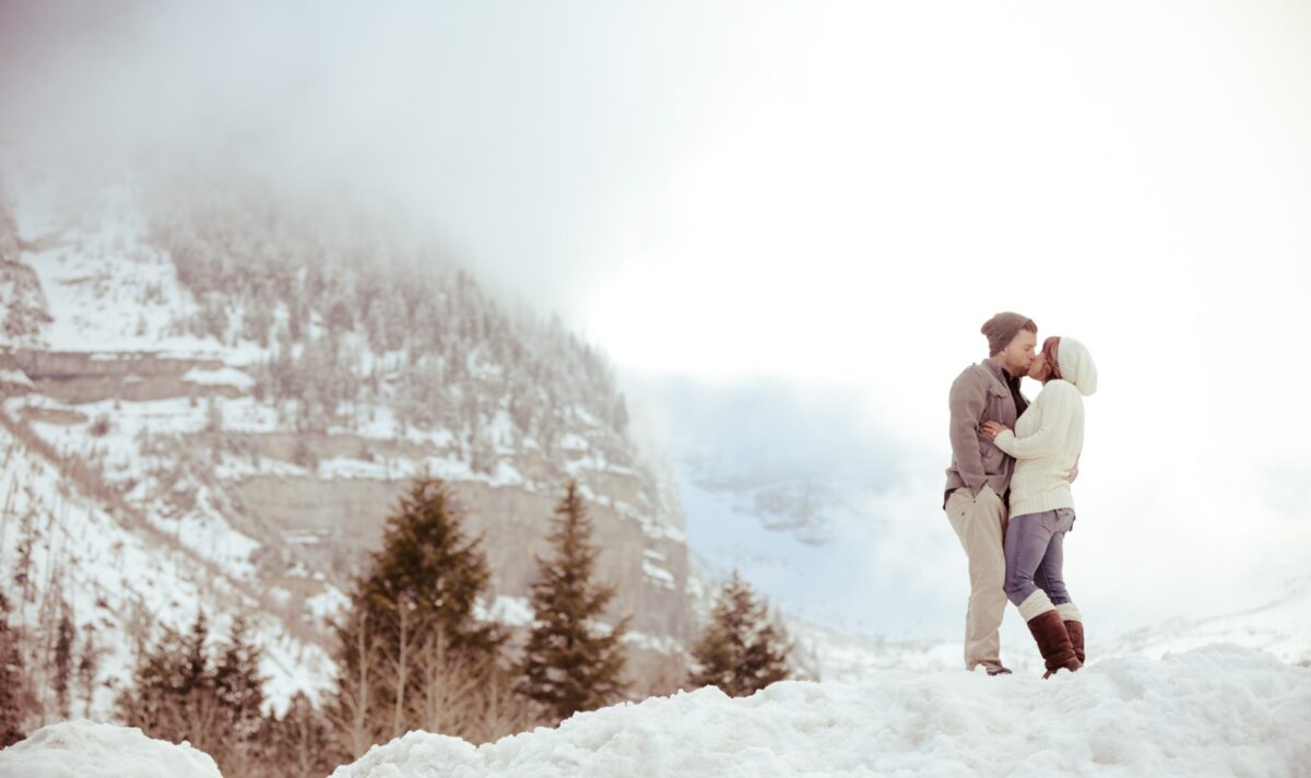 A man and woman kissing on a snowy mountaintop for their winter engagement photos | The Dating Divas