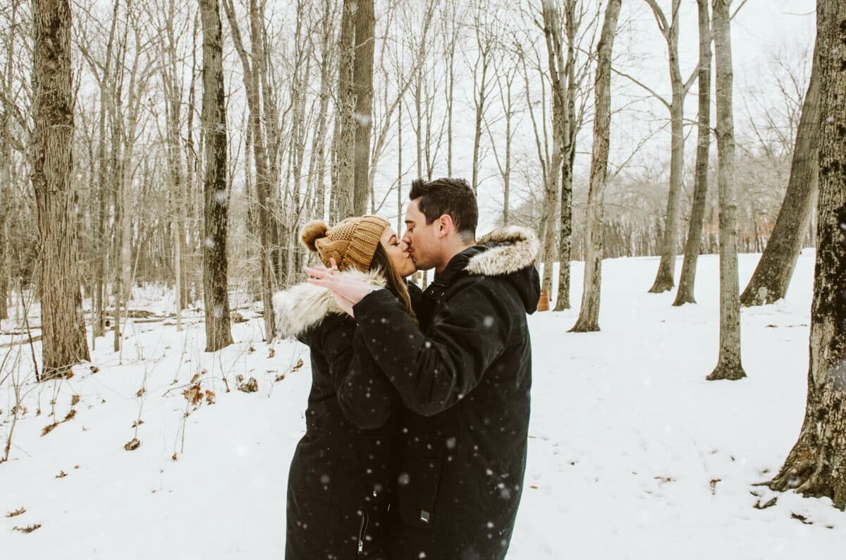 A couple kissing in a snowy forest for their winter engagement photos | The Dating Divas