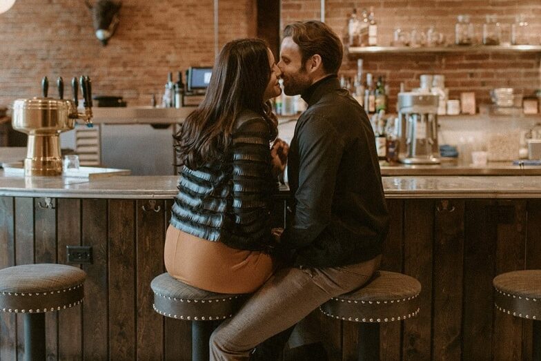 Engagement photo ideas showing a couple kissing while sitting on bar stools | The Dating Divas